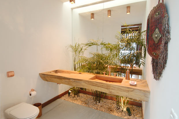 Nesta residência, o banheiro recebeu um canteiro com palmeiras localizado logo atrás da pia feita de madeira. A parede anterior é revestida por um espelho que duplica o visual do jardim. (Crédito: Landscape)<br>
