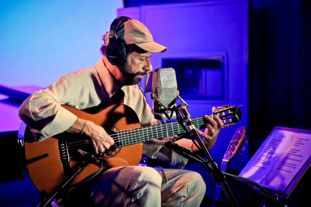 João Bosco, em estúdio, tocando violão, usando boné e fones de ouvido olhando para baixo