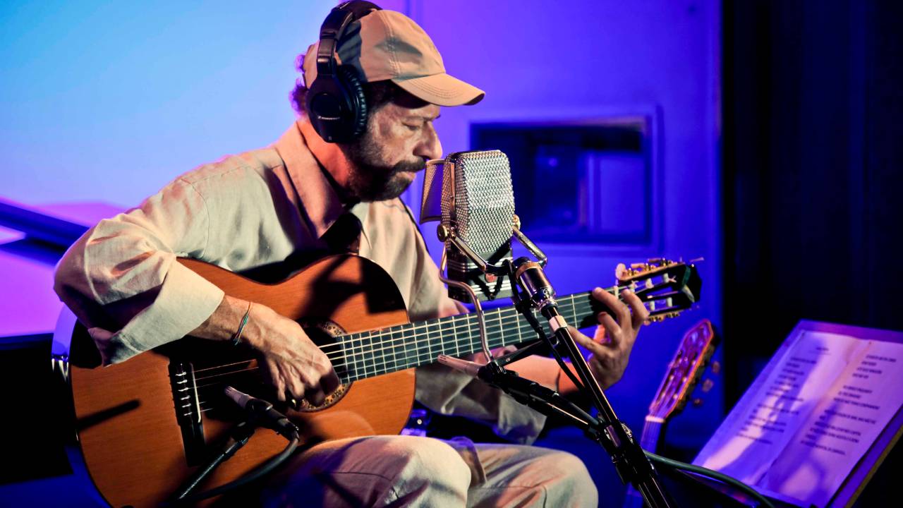 João Bosco, em estúdio, tocando violão, usando boné e fones de ouvido olhando para baixo