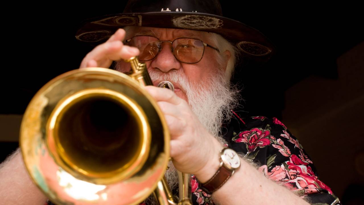 O músico Hermeto Pascoal, de chapéu e óculos de grau, toca um trompete, em primeiro plano