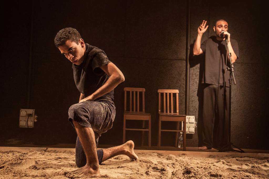 Dois atores em cena na peça Caranguejo Overdrive. Um deles está ajoelhado na areia e o outro ao fundo