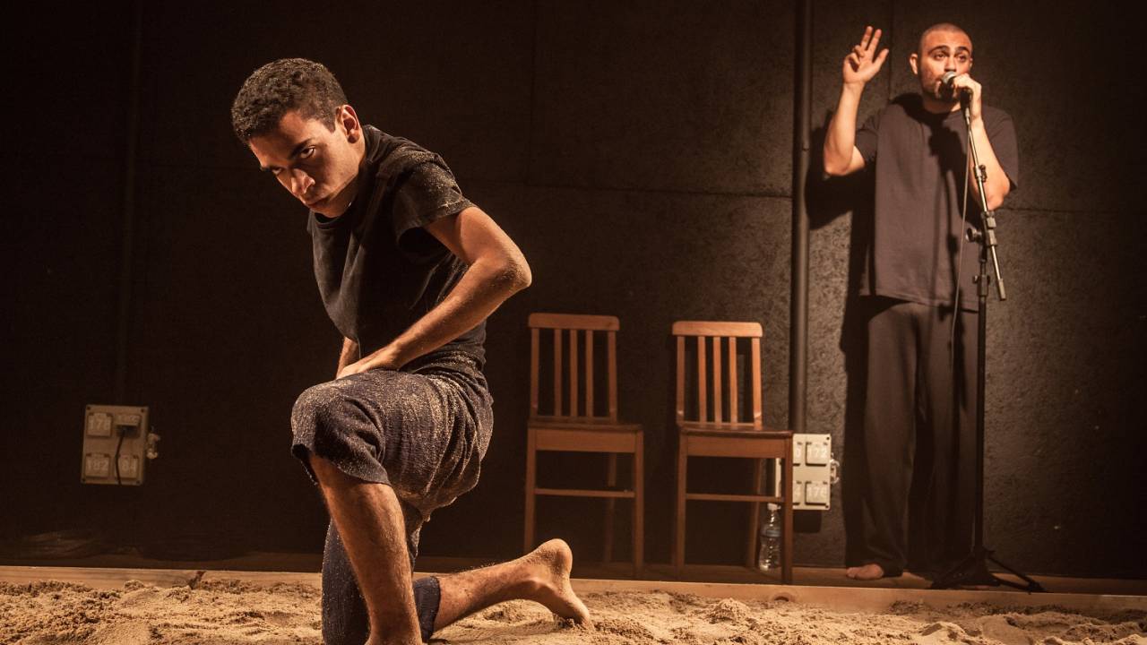 Dois atores em cena na peça Caranguejo Overdrive. Um deles está ajoelhado na areia e o outro ao fundo