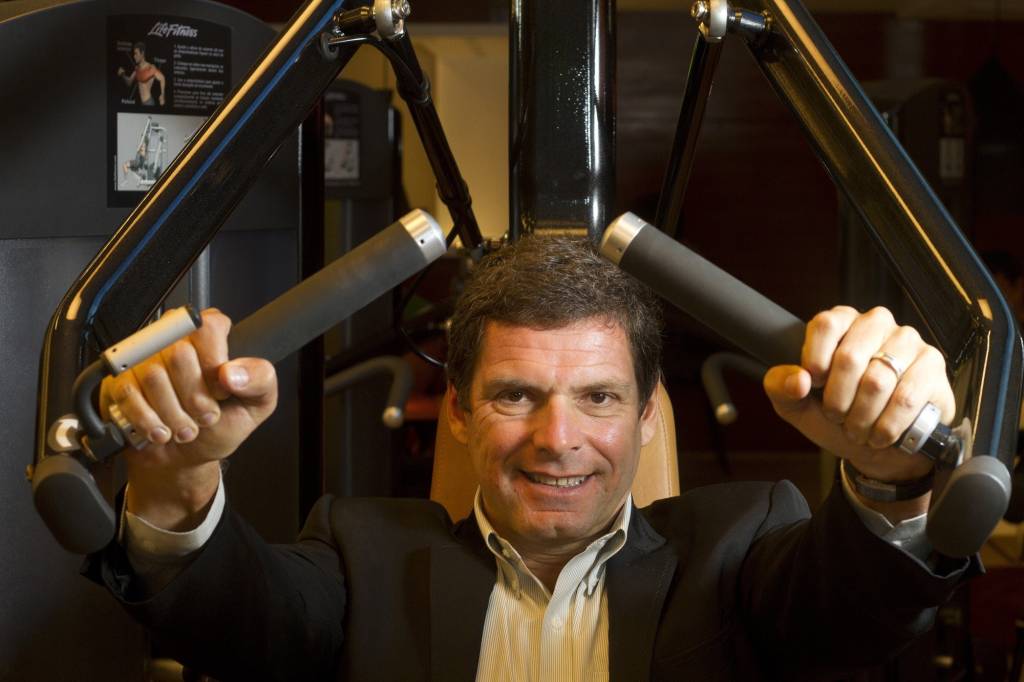 A imagem mostra o empresári Alexandre Accioly, de blazer, numa cadeira de fazer exercicios, numa academia de ginástica
