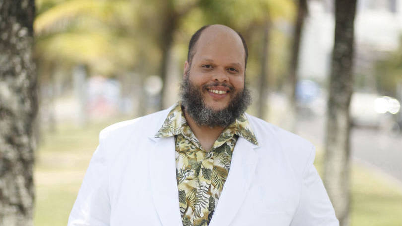 O cantor e compositor Ed Motta está sorrindo. Ele usa paletó branco e camisa florida.