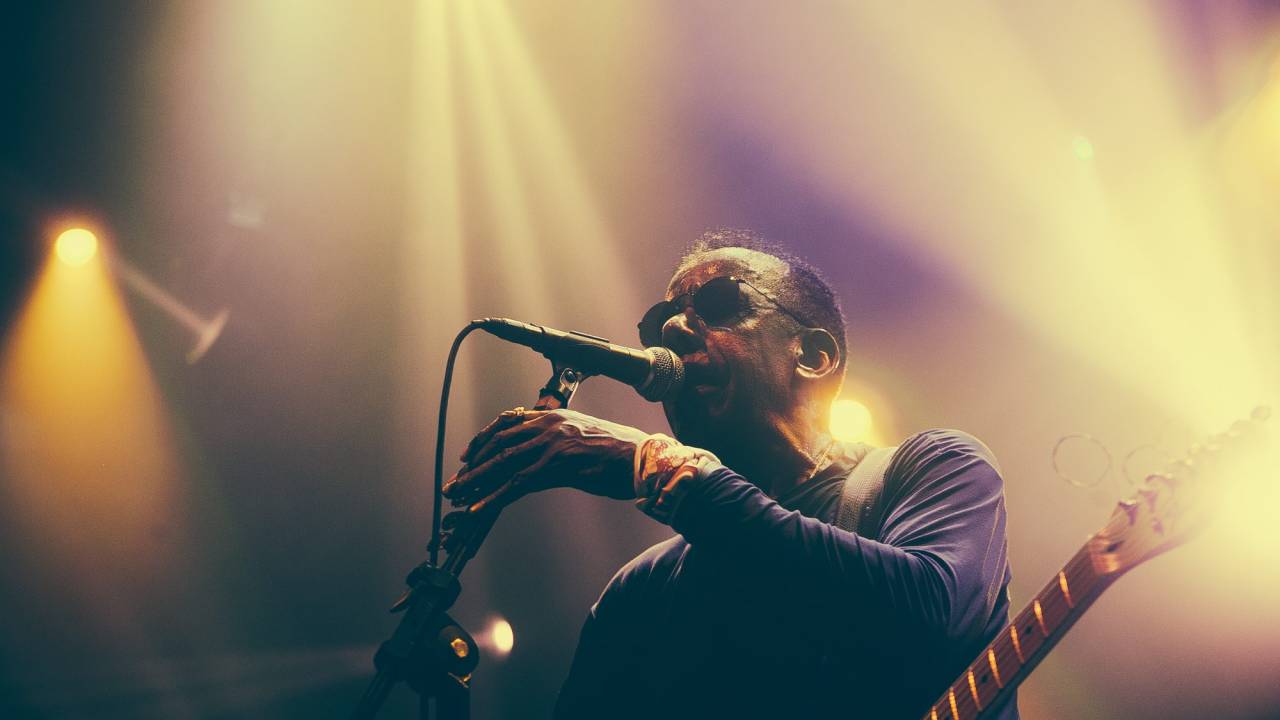 Jorge Ben Jor, de óculos escuros e blusa de manga comprida azul marinho, cantando.