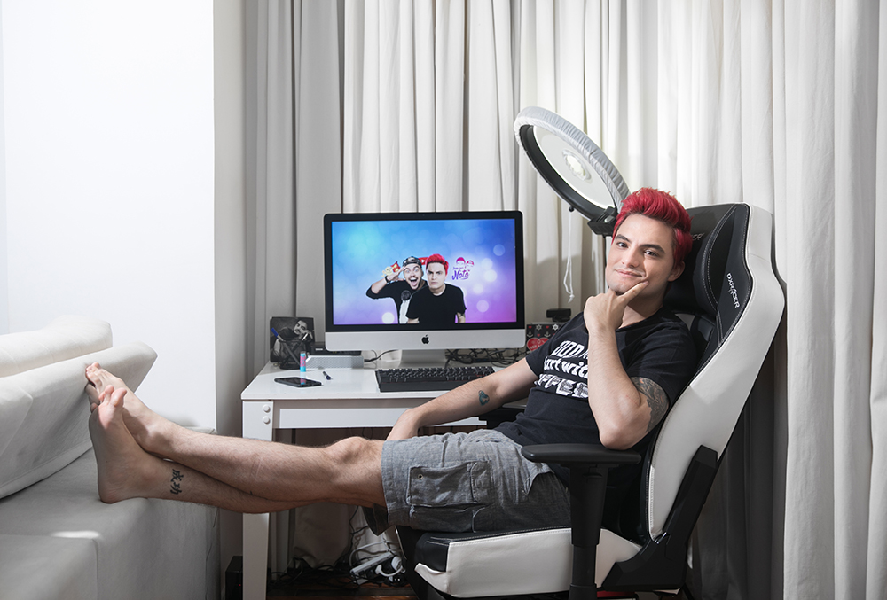 Foto mostra Felipe Neto, de cabelos vermelhos, sentado numa cadeira gamer, com a mão no queixo e um monitor e um ring light atrás dele