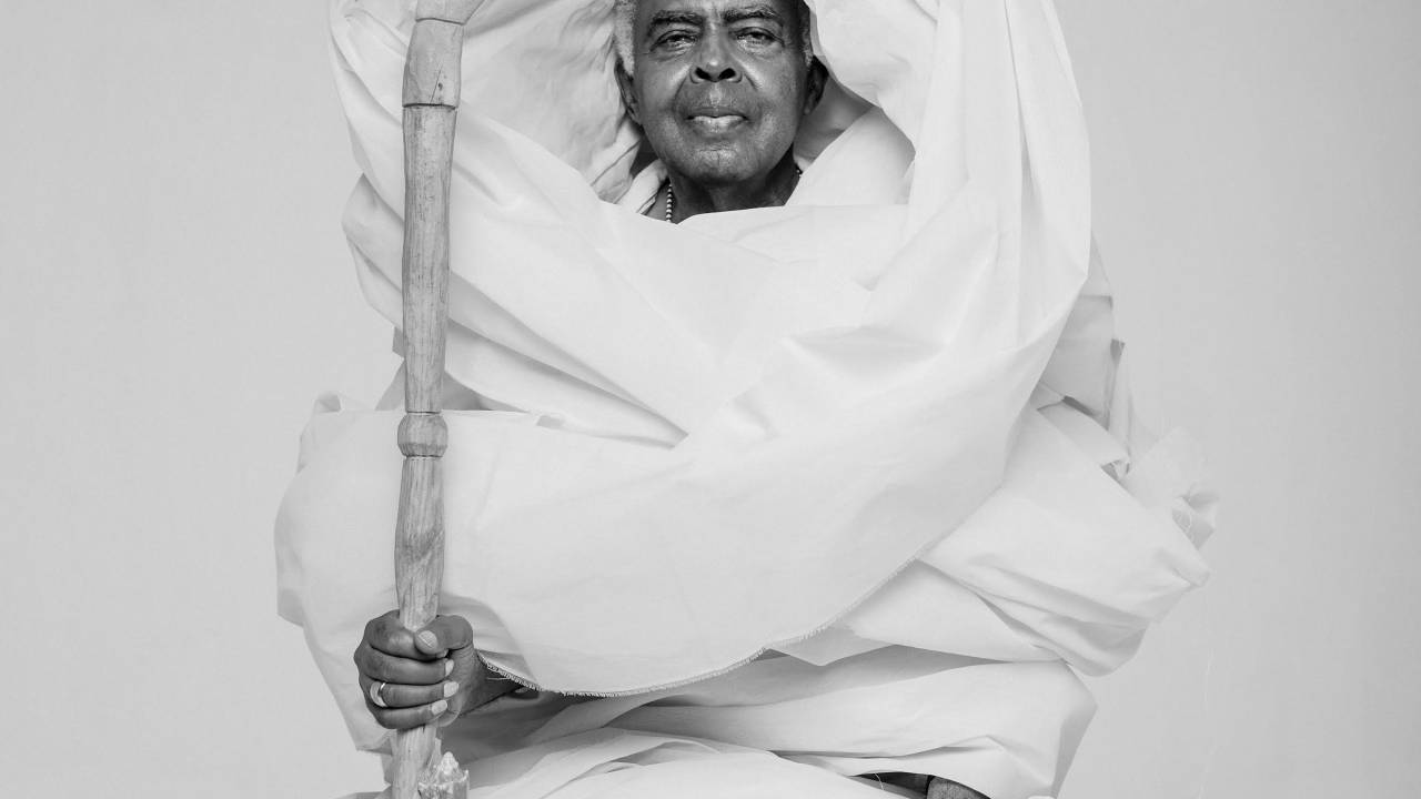 Gilberto Gil envolto por um pano branco e segurando um cajado