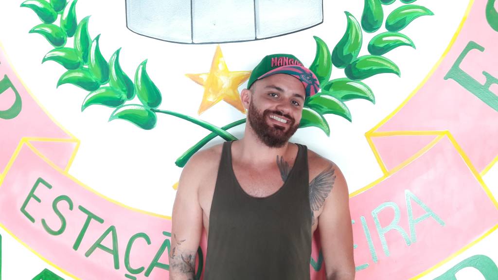 Carnavallesco da Mangueira sorri em frente a uma pintura do bandeira da escola