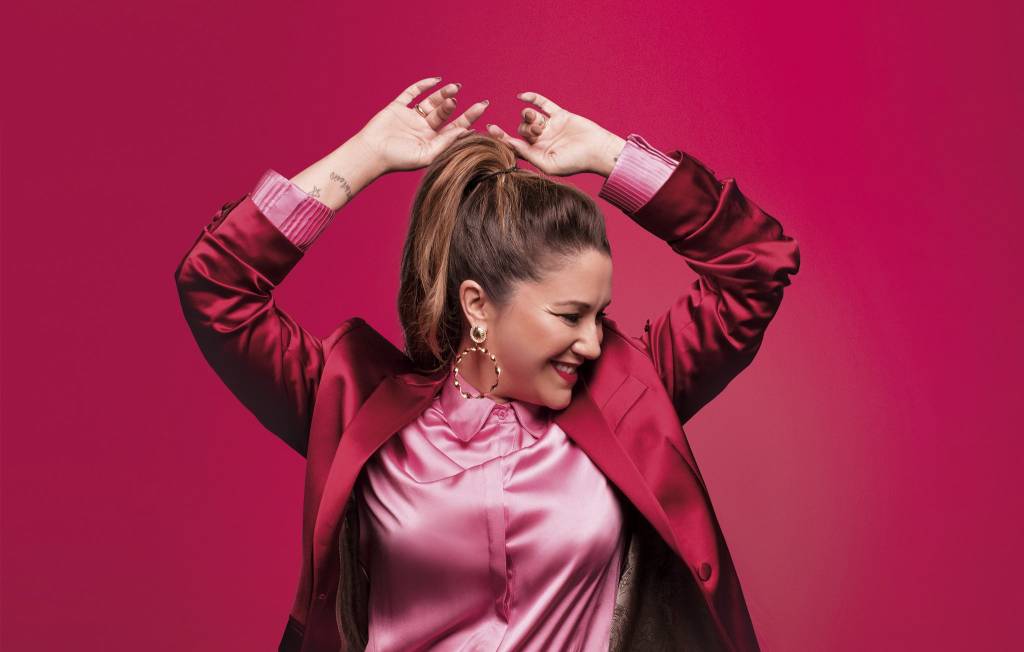 Maria Rita de terno vinho com camisa rosa sobre parede vinho. A cantora está com o rosto para o lado e os braços para o alto.