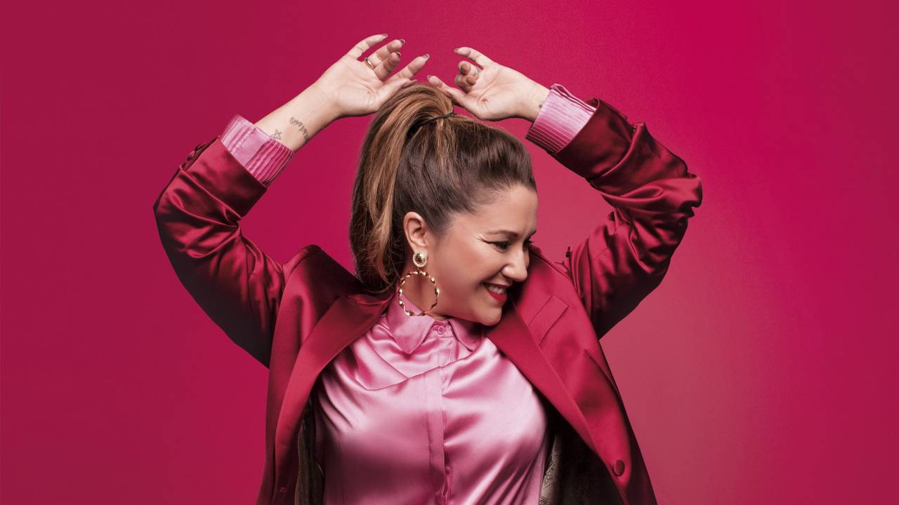 Maria Rita de terno vinho com camisa rosa sobre parede vinho. A cantora está com o rosto para o lado e os braços para o alto.