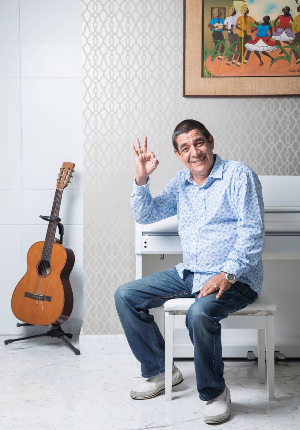 Zeca Pagodinho sentado num banco branco, com um violão ao lado dele, fazendo sinal de ok com a mão