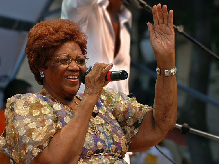 Dona Ivone Lara no palco, cantando, com uma das mãos para cima