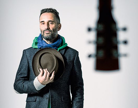 Jorge Drexler está em segundo plano usando paletó cinza, cachecóis coloridos e segurando um chapéu marrom