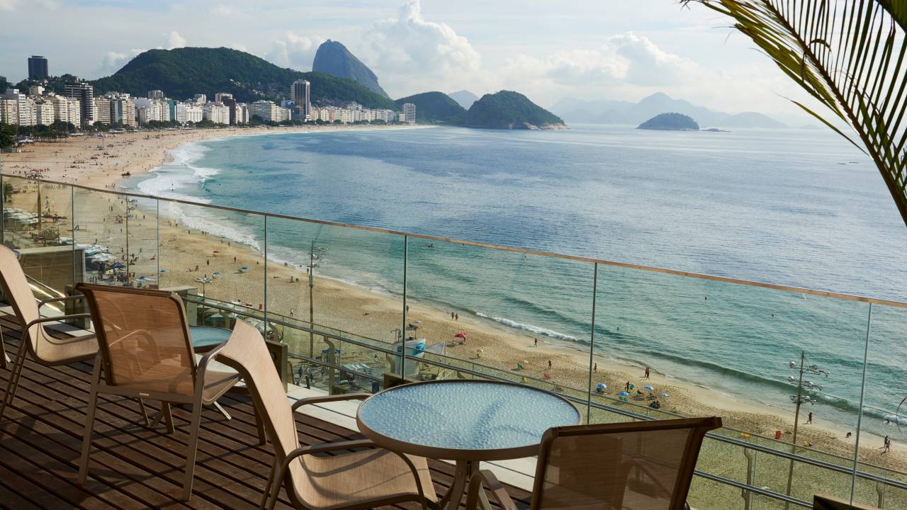 Uma mesa redonda com duas cadeiras em uma varanda de vidro do Grand Mercure Copacabana com vista para a praia