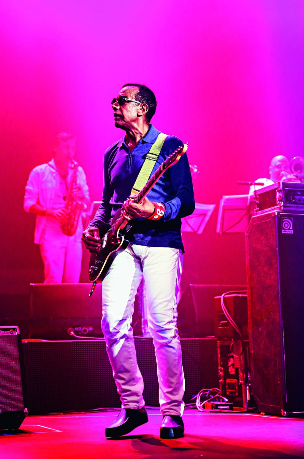 Jorge Ben Jor é um homem negro, está de camisa azul-escuro e calça branca, usando óculos de sol e tocando guitarra. A foto é iluminada por uma luz rosa.