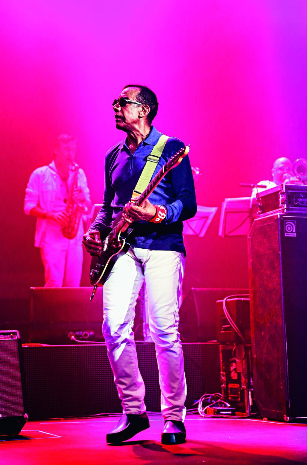 Jorge Ben Jor Jorge Ben Jor é um homem negro, está de camisa azul-escuro e calça branca, usando óculos de sol e tocando guitarra. A foto é iluminada por uma luz rosa.