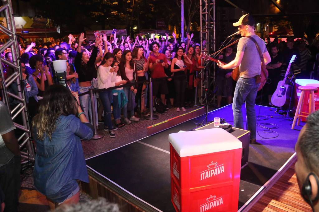 A imagem mostra o cantor nando reis no palco, visto de costas, com a plateia à sua frente
