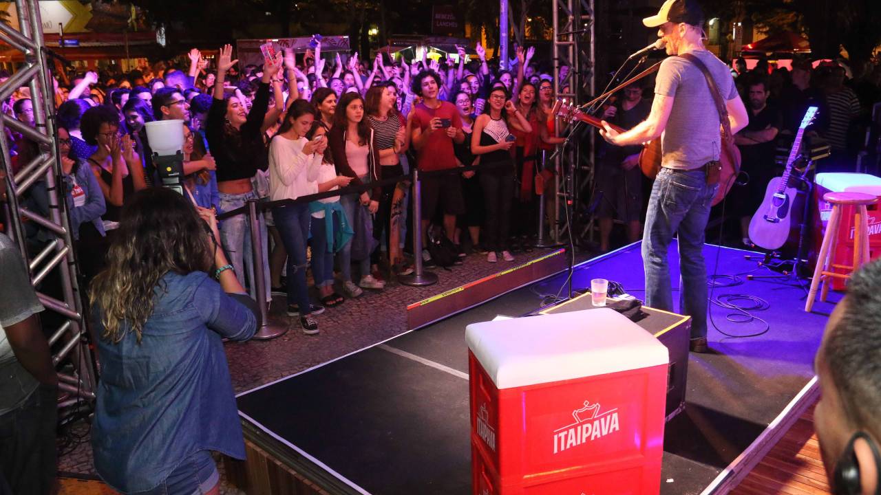 A imagem mostra o cantor nando reis no palco, visto de costas, com a plateia à sua frente