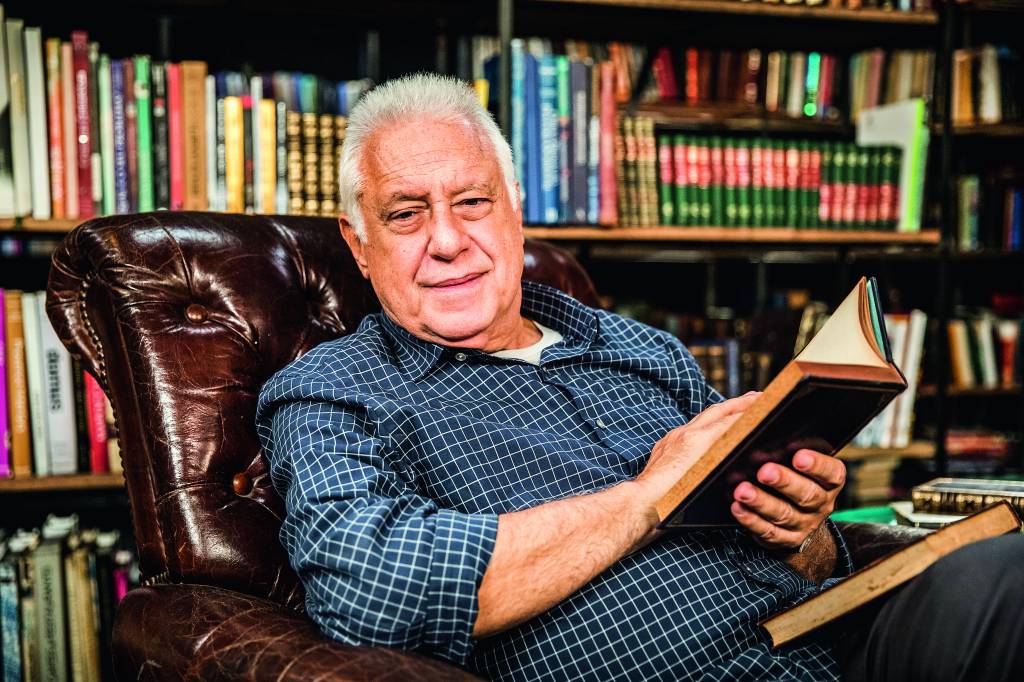 Antonio Fagundes na frente de uma estante de livros e segurando livro na mão