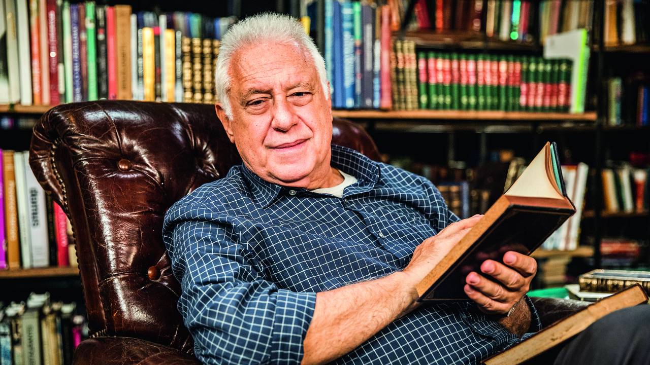 Antonio Fagundes na frente de uma estante de livros e segurando livro na mão