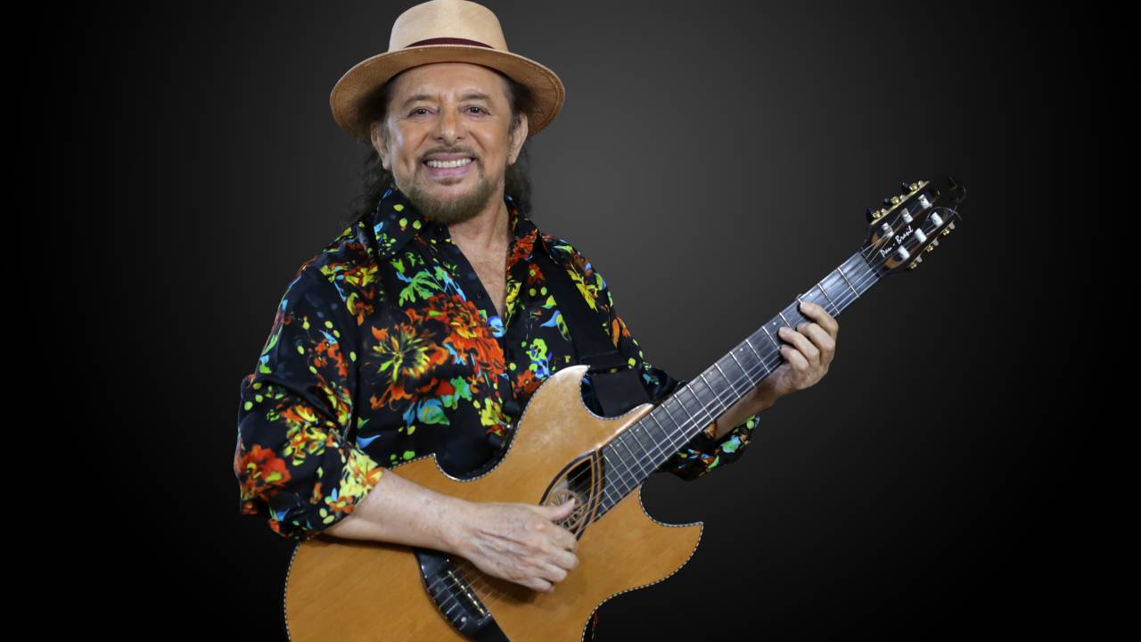 Geraldo Azevedo de chapéu e com violão nas mãos, sorrindo, com uma camisa colorida