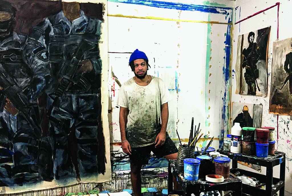 artista maxwell alexandre posa em seu atelier, há pincéis e telas espalhados pelo espaço