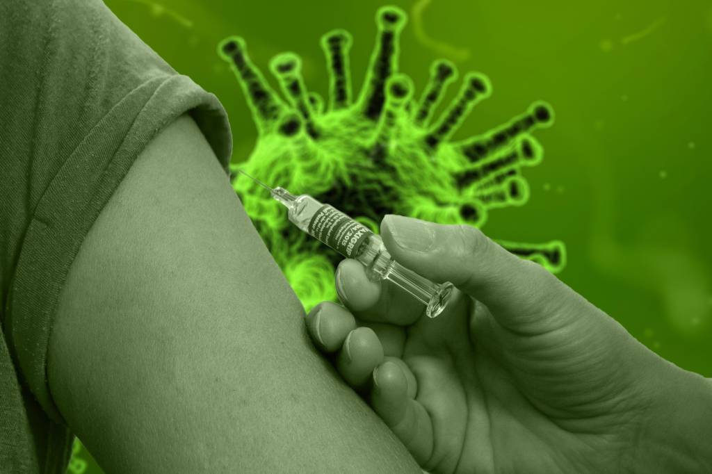 A imagem mostra um braço prestes a ser vacinado, e a ilustração do vírus ao fundo