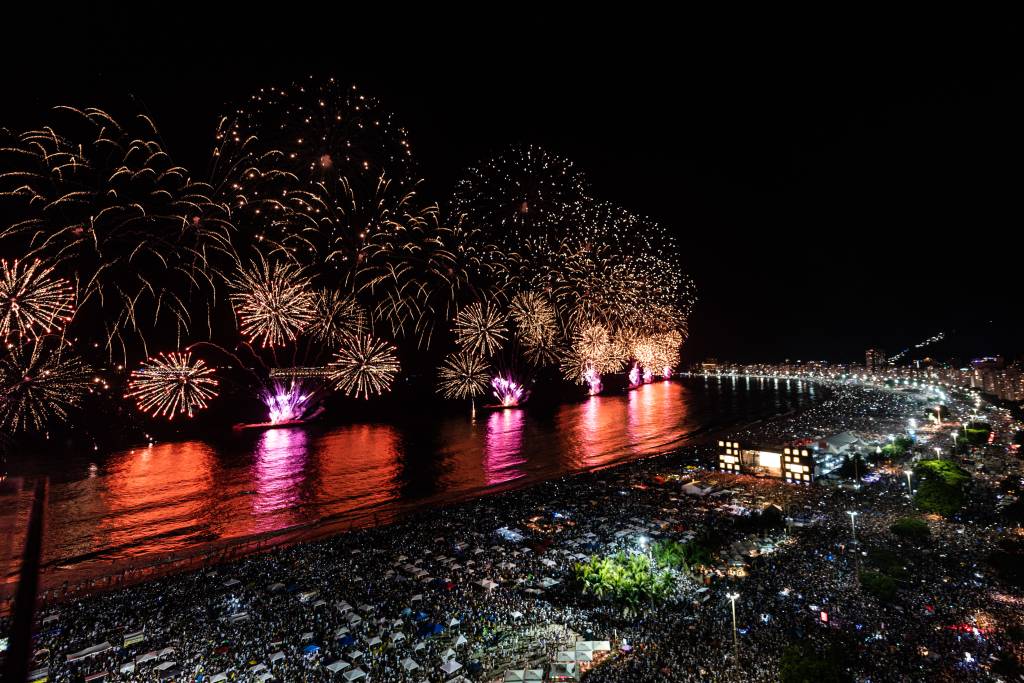 A imagem mostra a orla de Copacabana com fogos e multidão na areia na noite da virada