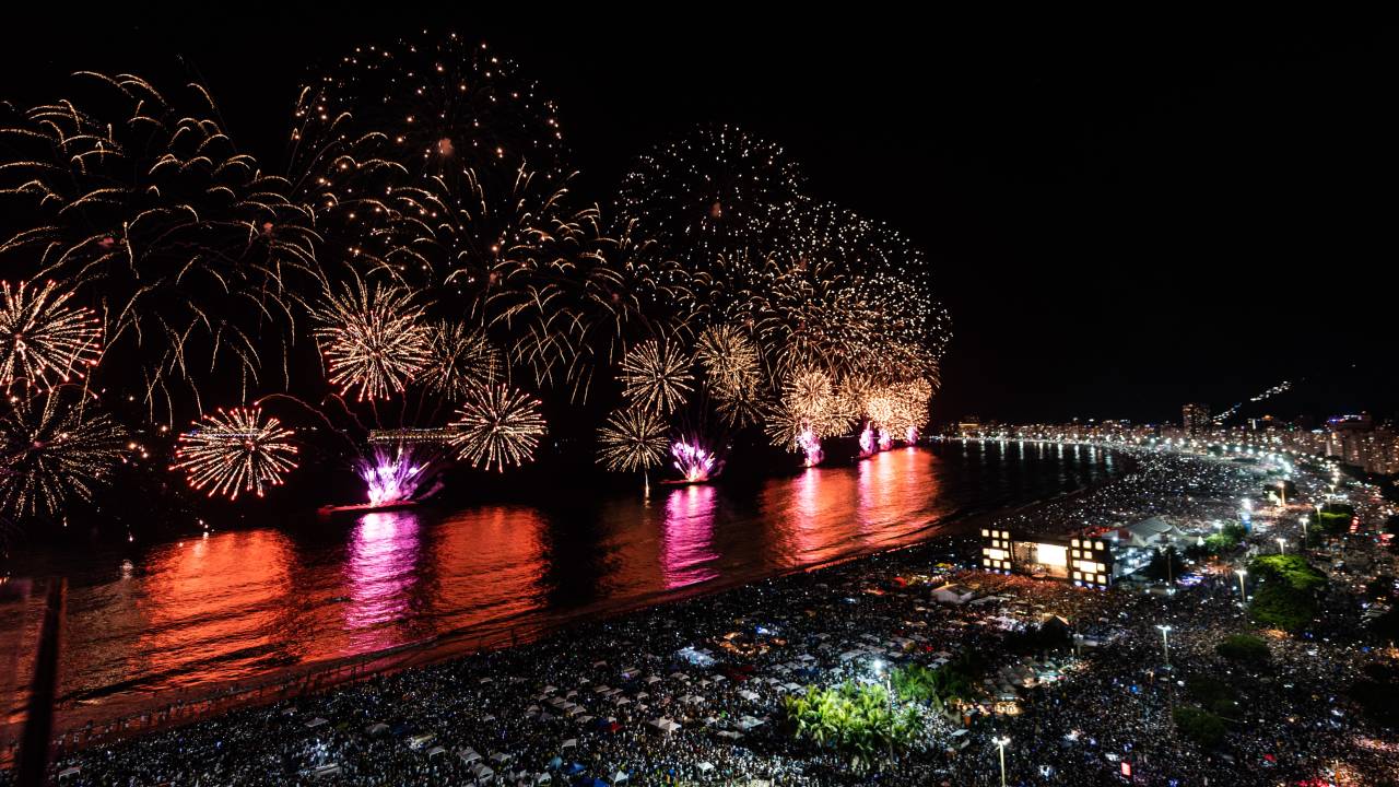 A imagem mostra a orla de Copacabana com fogos e multidão na areia na noite da virada