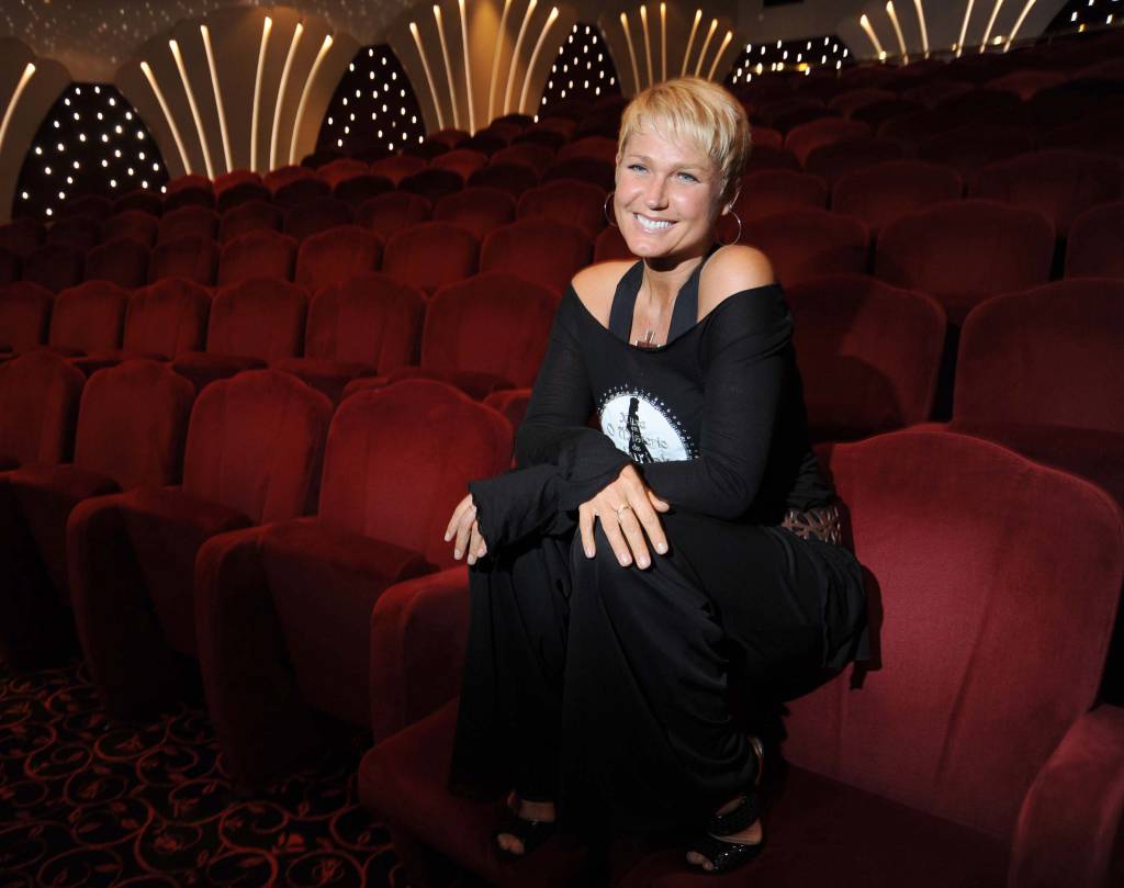 A imagem mostra a apresentadora Xuxa sentada na poltrona da plateia de um teatro, sorrindo para a câmera