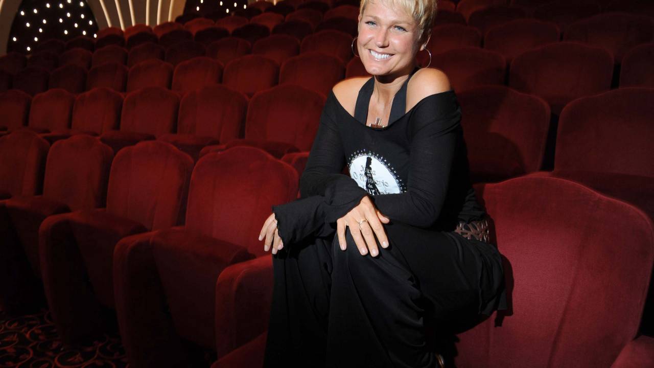 A imagem mostra a apresentadora Xuxa sentada na poltrona da plateia de um teatro, sorrindo para a câmera