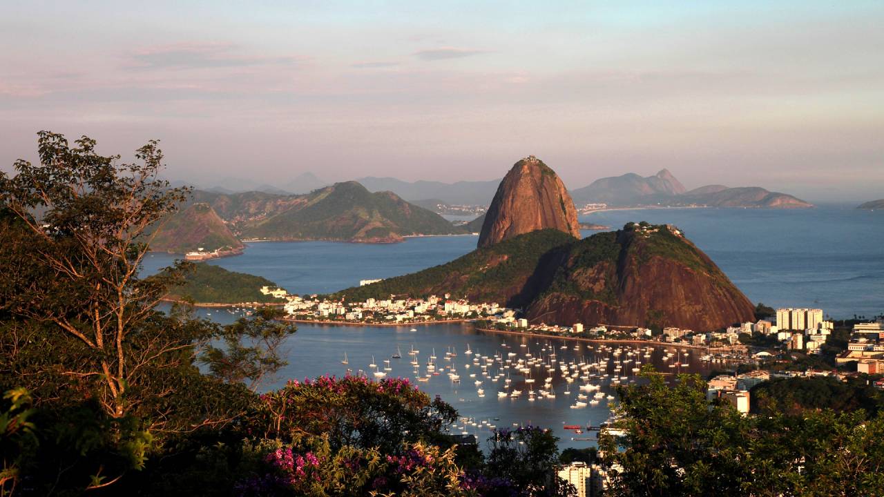 A imagem mostra o Rio de Janeiro do alto, com o Morro da Urca e o Pão de Açúcar, além da Baía de Guanabara