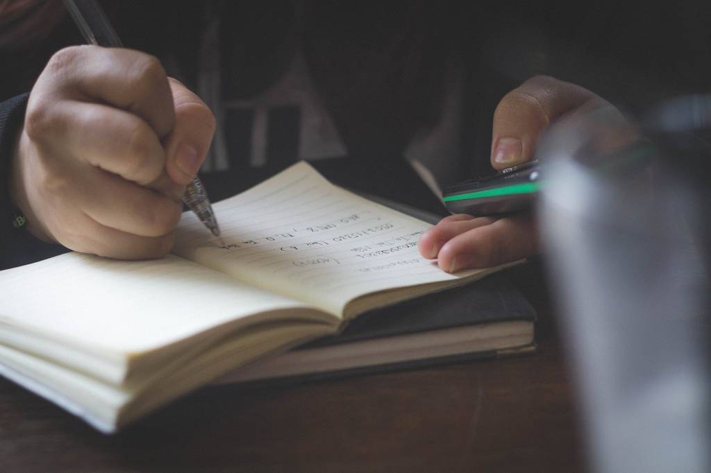A imagem mostra uma pessoa escrevendo num caderno com um celular na outra mão