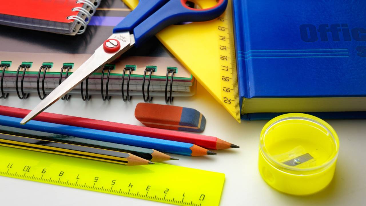 mesa com materiais escolares espalhados, como tesoura, lápis, borrancha, caderno e régua
