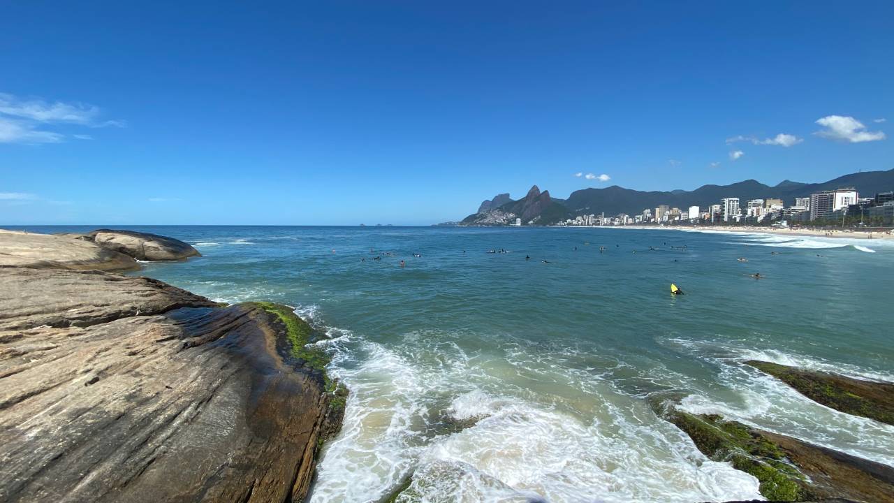 A imagem mostra um dia de céu azul na Praia de Ipanema, sob o ponto de vista de quem está no Arpoador