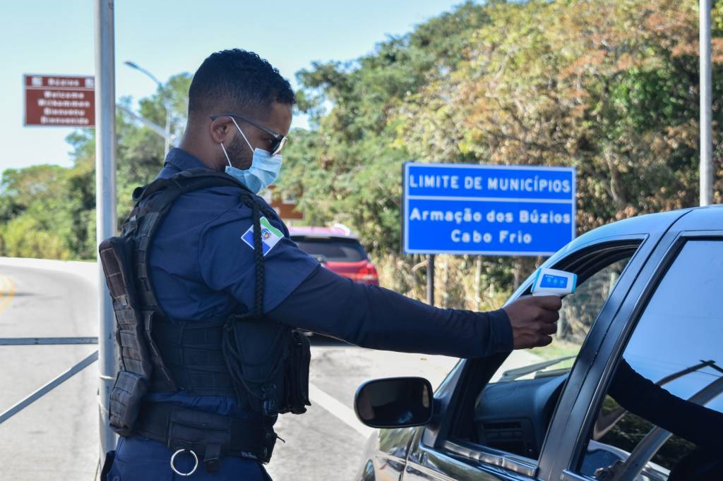 A imagem mostra um policial aferindo a temperatura de um visitante, que está dentro do carro, na chegada à cidade de Búzios