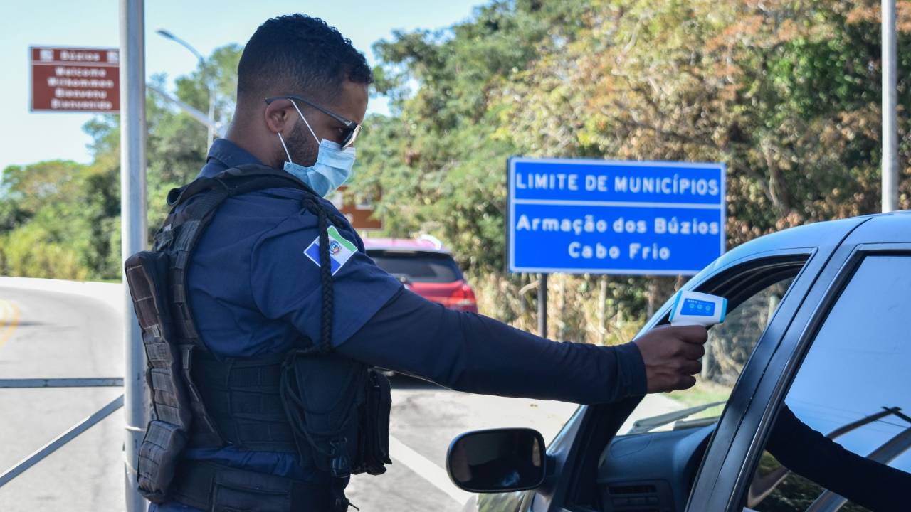 A imagem mostra um policial aferindo a temperatura de um visitante, que está dentro do carro, na chegada à cidade de Búzios