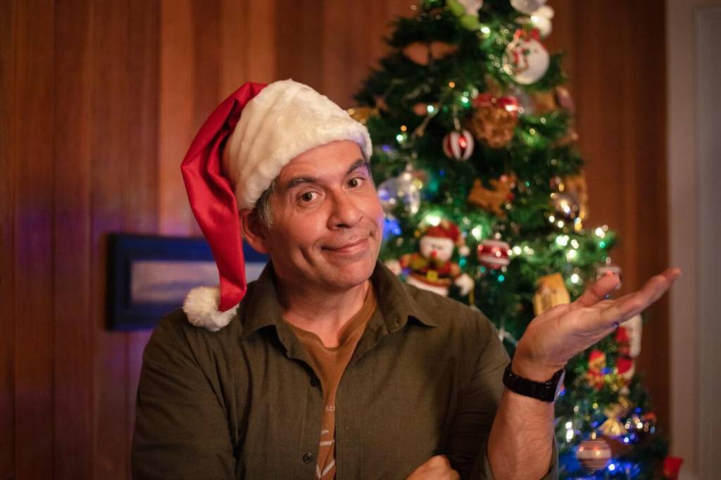 Leandro Hassum, com gorro de Papai Noel, sorrindo sem mostrar os dentes. Há uma árvore de Natal ao fundo