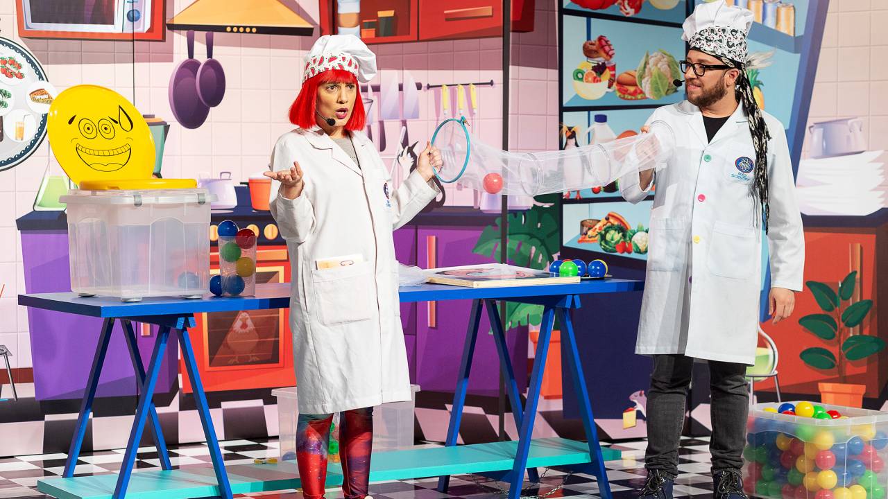 Dois atores, com jaleco de cientistas, num cenário de cozinha multicolorido