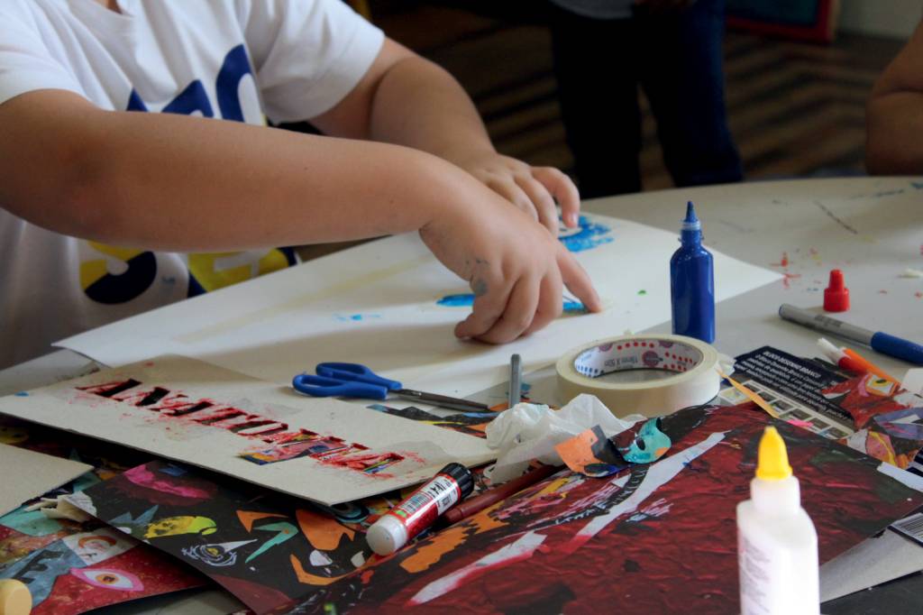 braços de criança fazendo pintura. papéis coloridos, cola e tinta espalhados pela mesa