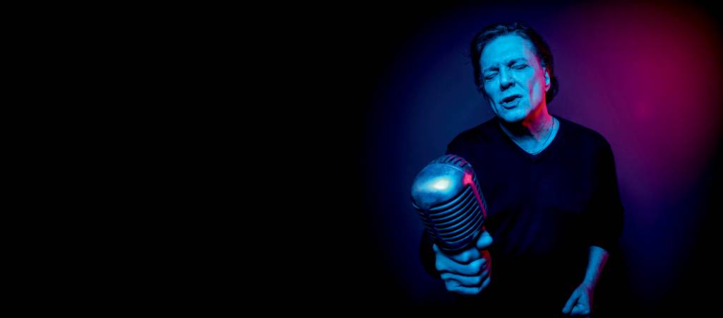 Fabio Jr num fundo escuro e bem azulado, segurando um microfone, de olhos fechados, cantando