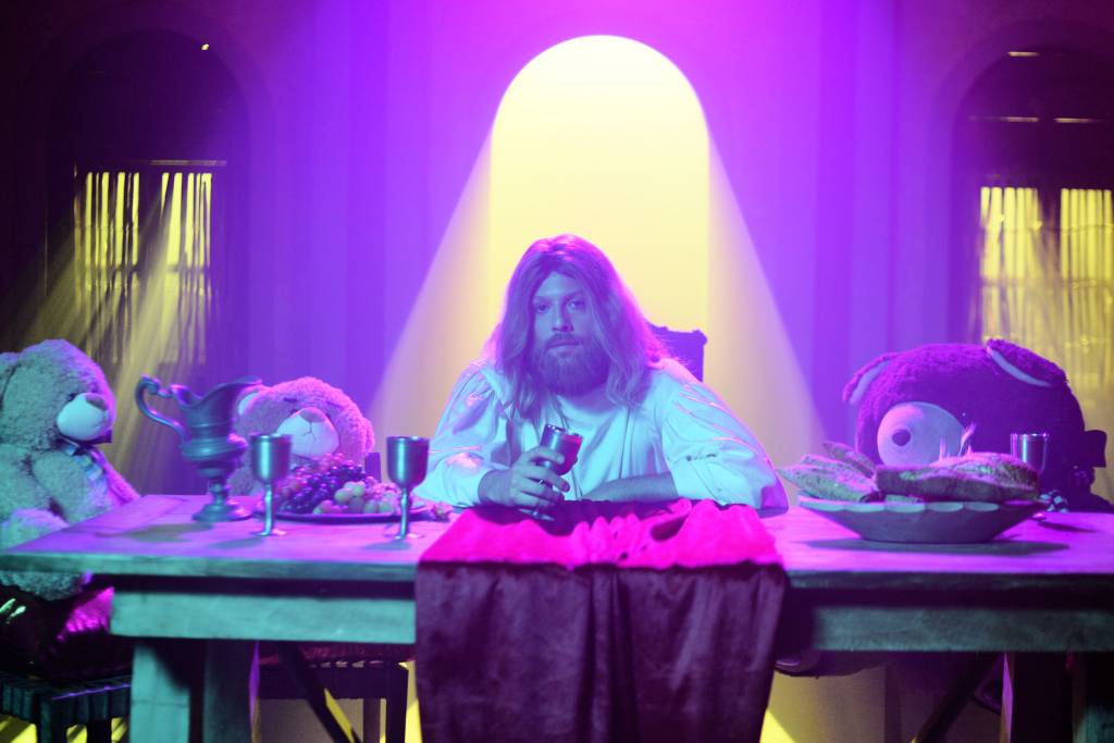 Jesus à mesa, ao lado de bichinhos de pelúcia. Há uma iluminação arroxeada
