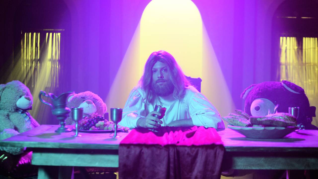 Jesus à mesa, ao lado de bichinhos de pelúcia. Há uma iluminação arroxeada