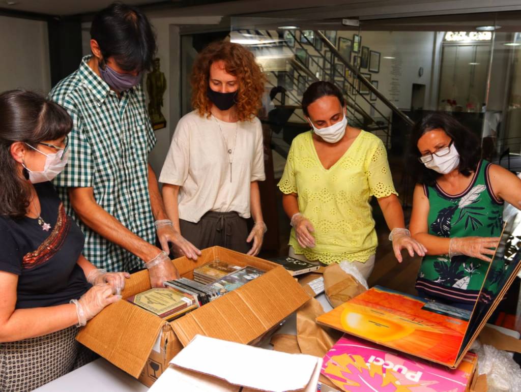 Cinco pessoas, de máscaras, analisam objetos como livros e discos dispostos em cima de uma mesa