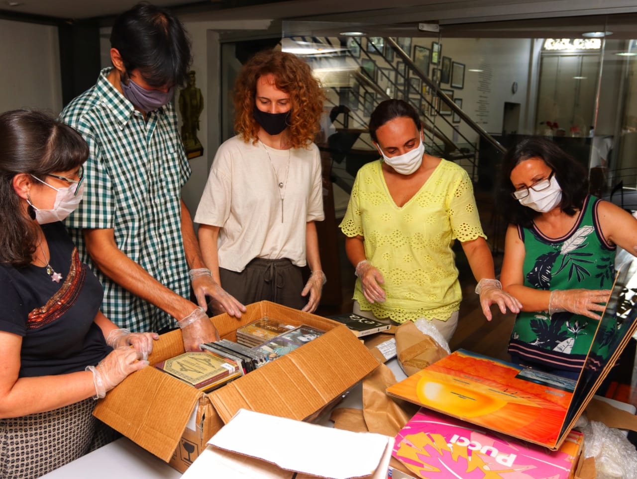 Museu da Imagem e do Som do Rio recebe o acervo de José Wilker (2) Cinco pessoas, de máscaras, analisam objetos como livros e discos dispostos em cima de uma mesa