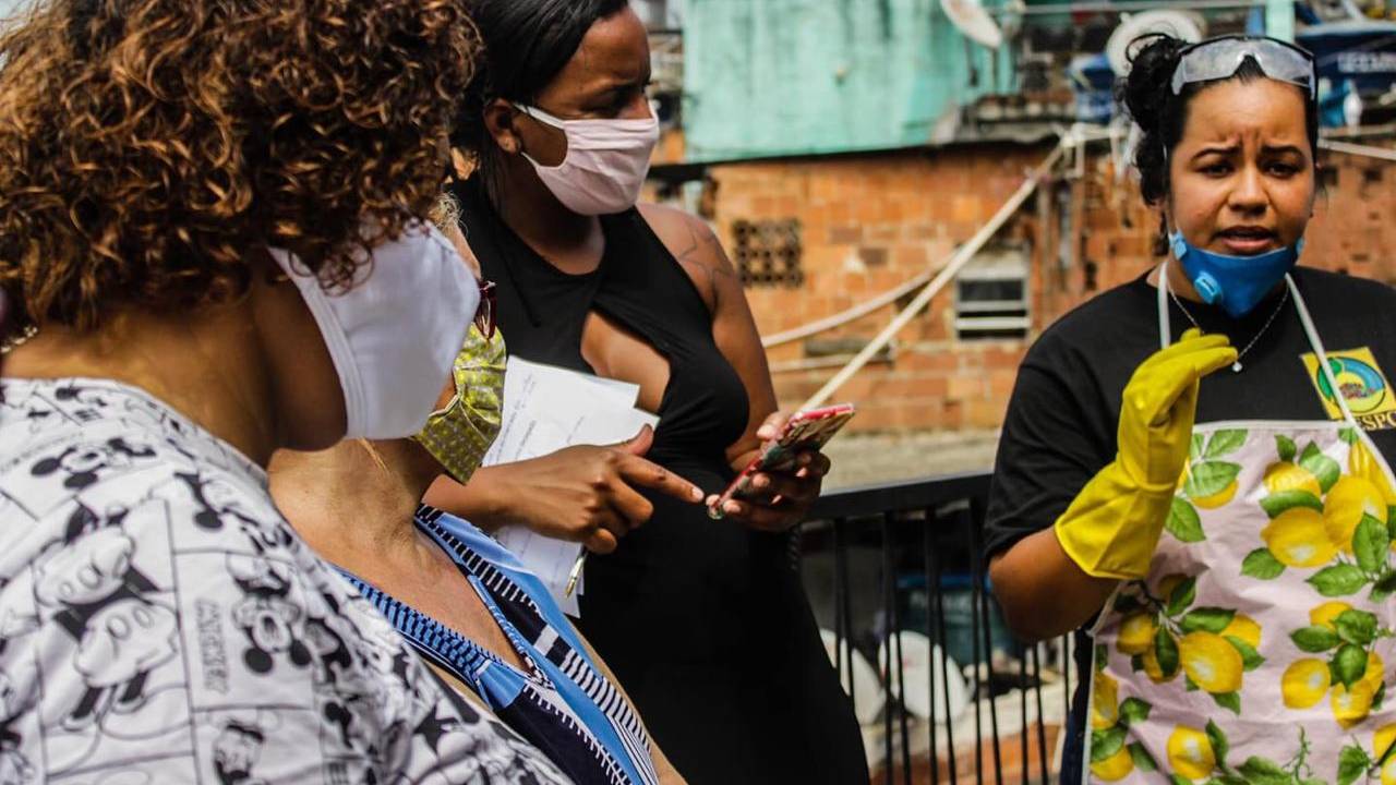 Três mulheres de máscara ensinam a fazer sabão líquido na favela