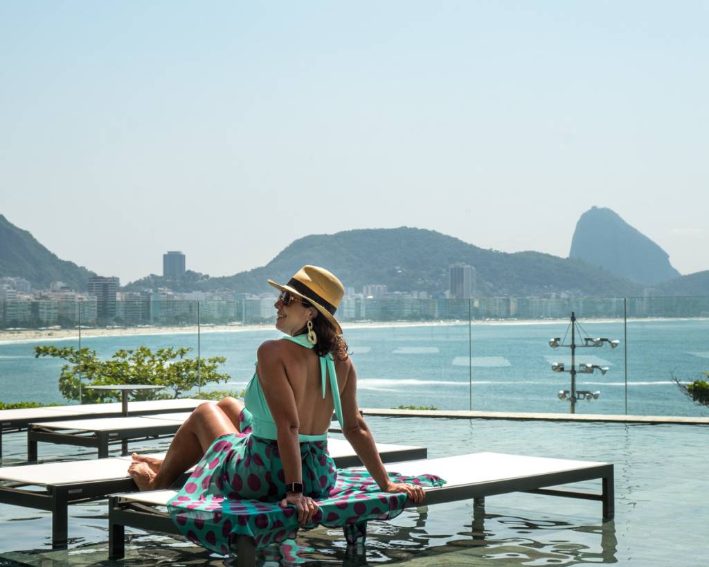 mulher de maiô e chapé panamá sentada em espreguiçadeira na beira da piscina. Vista para a Praia de Copacabana