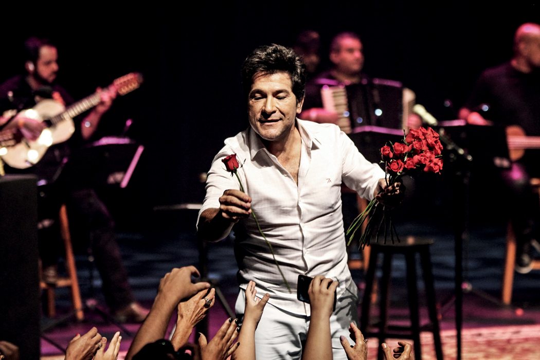 Cantor Daniel, vestido de branco, no palco, joga flores para a plateia. É possível ver, ao fundo, músicos que o acompanham