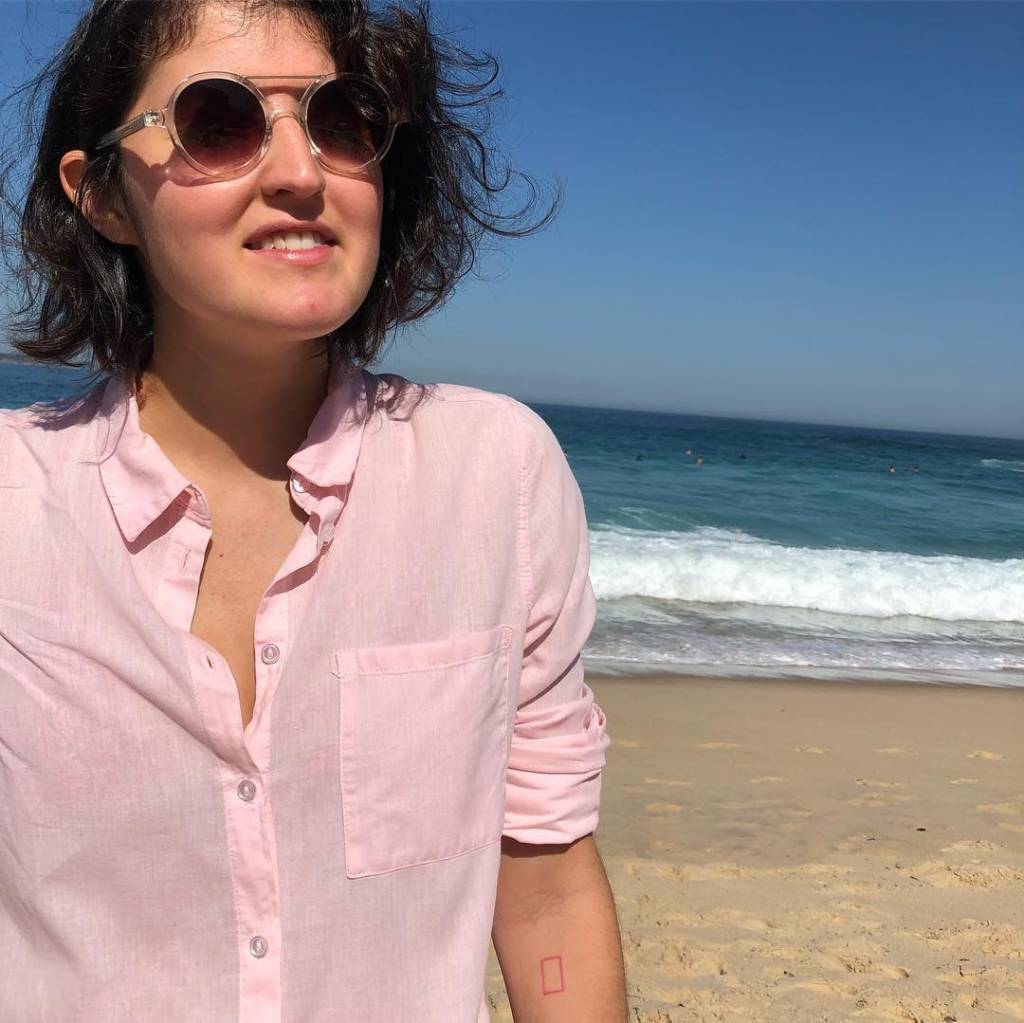 Gabriela Davies está na praia, vestido uma camisa rosa e óculos escuros
