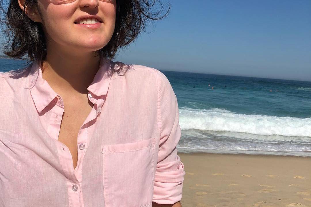 Gabriela Davies está na praia, vestido uma camisa rosa e óculos escuros